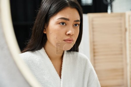 mujer asiática triste con pelo morena examinando su cara con acné en el espejo del baño, problema de la piel