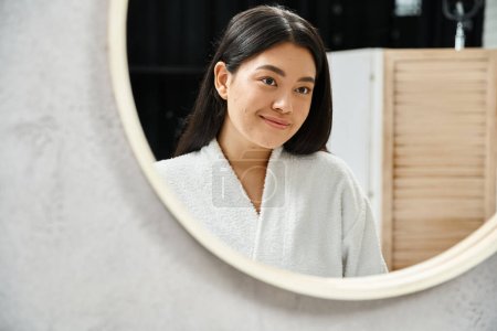 happy asian girl with brunette hair examining her face with acne in bathroom mirror, skin issue