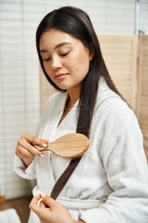 young asian woman in robe combing her brunette healthy hair with wooden hairbrush in bathroom