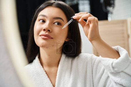 Foto de Joven y morena mujer asiática en bata de baño aplicando suero facial para tratar el acné en la cara cerca del espejo - Imagen libre de derechos