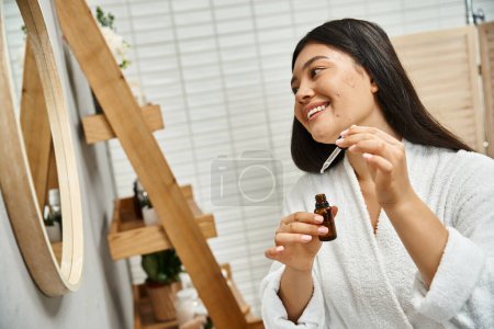 cheerful and young asian woman in bath robe applying facial serum to treat acne on face near mirror