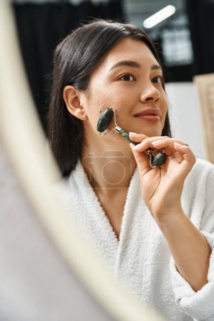 smiling asian woman with brunette hair using jade roller for a facial massage in bathroom, skin care