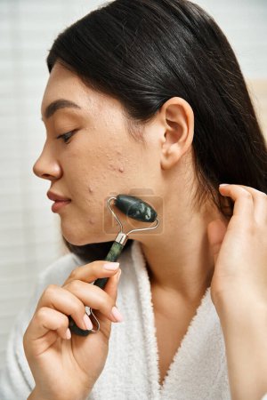 close up photo of young asian woman with acne doing face massage with jade roller in bathroom
