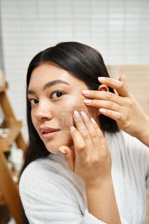 junge und brünette asiatische Mädchen im Bademantel knallen Pickel auf ihr Gesicht, Hautprobleme oder echte Menschen