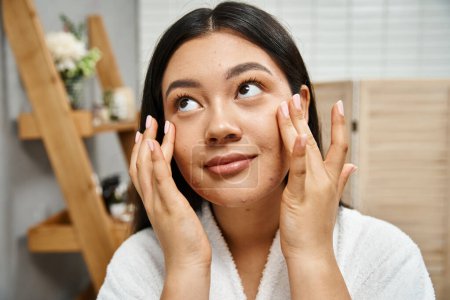 Hautpflegekonzept, junge Asiatin im Bademantel, die Feuchtigkeitscreme auf das Gesicht aufträgt und nach oben schaut