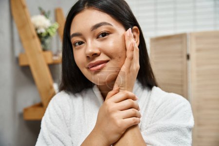 portrait of happy young asian woman with acne touching cheek and looking at camera, skin condition