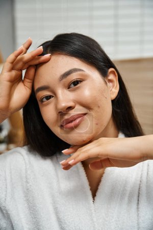 portrait of happy young asian woman with acne touching face and looking at camera, skin care