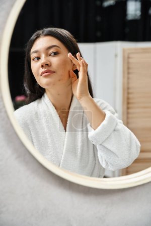 Photo for Young asian woman with acne applying cream on face and looking at mirror in bathroom, skin issues - Royalty Free Image