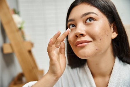 brunette asian woman with acne applying cream on face and looking up in bathroom, skin issues