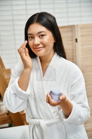 happy asian woman with acne applying cream on face and smiling while looking at camera in bathroom