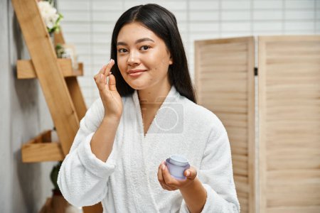 Photo for Cheerful asian woman with acne applying cream on face and smiling while looking at camera at home - Royalty Free Image