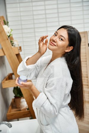 Photo for Happy and pretty asian woman with acne applying cream on face and smiling while looking at camera - Royalty Free Image