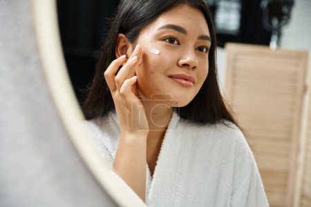 Foto von jungen asiatischen Frau mit brünetten Haaren Anwendung Akne-Behandlung Creme und Blick auf den Spiegel