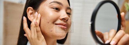 junge und brünette asiatische Frau mit Pickeln untersucht ihr Gesicht im Spiegel, Hautprobleme Banner