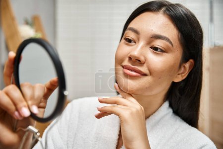 young and smiling asian woman with pimples examining her face in mirror, skin issues of real people