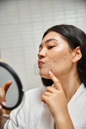 joven asiático mujer con morena cabello y espinillas examinando su cara en espejo, problemas de piel