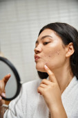young asian woman with brunette hair and pimples examining her face in mirror, skin issues magic mug #692759472