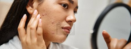 young asian woman with brunette hair and pimples looking at her face in mirror, skin issues banner