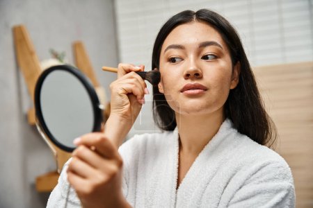 mujer asiática joven con cabello moreno y acné aplicando polvo con cepillo cosmético, problemas de piel