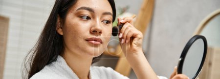 young asian woman with brunette hair applying makeup over acne-prone skin with brush, banner