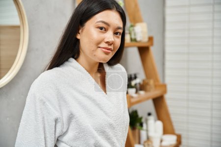 portrait of young asian woman in bath robe standing in modern bathroom and looking at camera