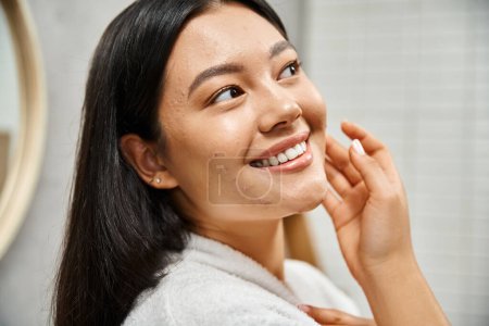 Foto de Primer plano de feliz joven asiático con acné propenso a la piel de pie en moderno cuarto de baño y mirando a la cámara - Imagen libre de derechos