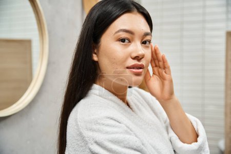 brunette young asian with acne-prone skin standing in modern bathroom and looking at camera