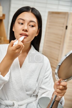 jeune et brune asiatique femme en peignoir de bain appliquer baume à lèvres et regarder miroir dans la salle de bain