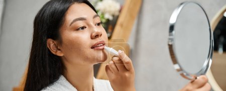 junge und brünette asiatische Frau im Bademantel Anwendung Lippenbalsam und Blick auf Spiegel, Banner