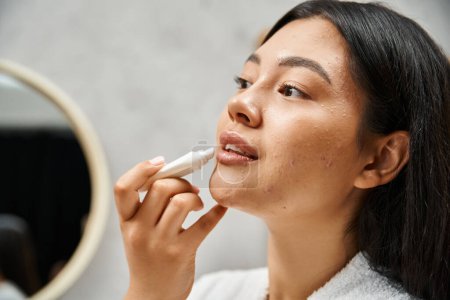 junge und brünette asiatische Frau mit Akne Anwendung Lippenbalsam und Blick auf Spiegel im Badezimmer