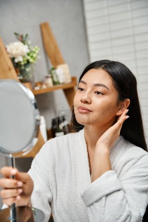 Porträt einer jungen Asiatin im Mantel, die zu Akne neigende Haut berührt und im Badezimmer in den Spiegel schaut