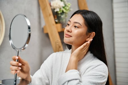 brünette und junge asiatische Frau mit Akne-anfälliger Haut beim Blick in den Spiegel im modernen Badezimmer, Hautpflege