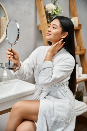 Porträt einer brünetten und jungen asiatischen Frau mit zu Akne neigender Haut, die im modernen Badezimmer in den Spiegel schaut