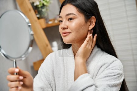 Porträt einer brünetten und jungen asiatischen Frau mit Makeln beim Anblick eines Spiegels im modernen Badezimmer