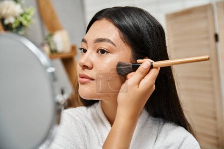 young asian woman with brunette hair and acne applying face powder and looking at mirror, skin issue