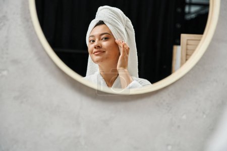 Photo for Young asian woman with white towel on head touching acne prone skin and looking at bathroom mirror - Royalty Free Image
