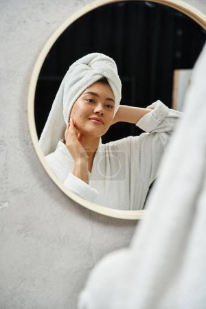 mujer asiática joven con toalla blanca en la cabeza y piel propensa al acné mirando el espejo del baño en casa