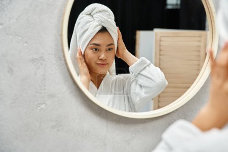 junge Asiatin mit Handtuch auf dem Kopf und zu Akne neigender Haut beim Blick auf den Badezimmerspiegel zu Hause
