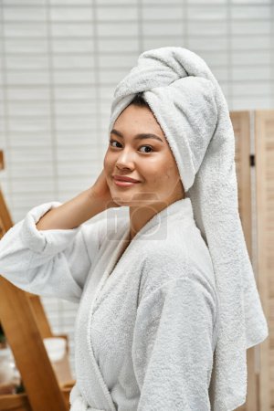 cheerful and young asian woman in robe with white towel on head looking at camera in bathroom