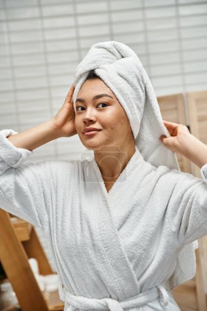 young asian woman in robe with white towel on head looking at camera in bathroom, skin issues