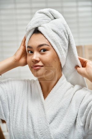 bonita mujer asiática en bata con toalla blanca en la cabeza mirando a la cámara en el baño, problemas de piel