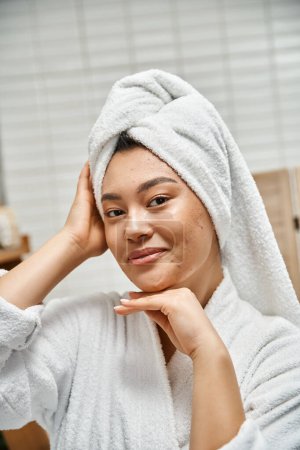joyful asian woman with acne and white towel on head looking at camera in bathroom, skin issues