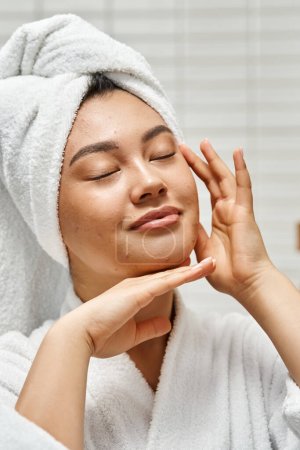Foto de Mujer asiática alegre con acné y toalla blanca en la cabeza de pie con los ojos cerrados en el baño, vertical - Imagen libre de derechos