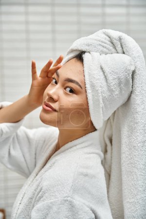 young asian woman with acne prone skin with towel on head posing in bathroom at home, vertical