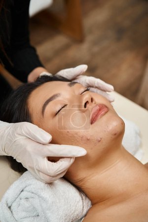 facial massage for acne-prone skin, masseuse in latex gloves and asian woman with closed eyes