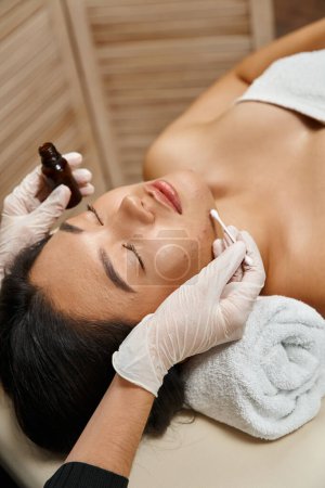 Hautpflege, Therapeut appliziert Behandlungsserum mit Wattestäbchen auf asiatische Frau mit Akne-anfälliger Haut