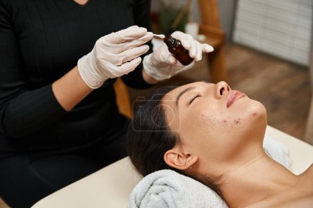 Therapeutin wendet Behandlungsserum mit Wattestäbchen auf Gesicht asiatischer Frauen mit Akne-anfälliger Haut an