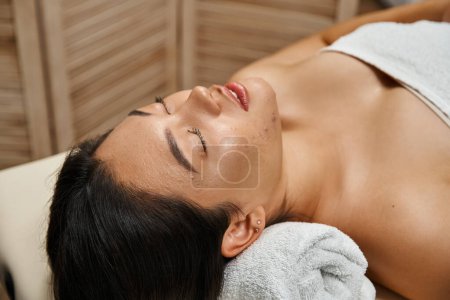 young and brunette asian woman with closed eyes lying in wellness center, spa and facial treatment