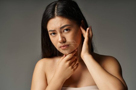 worried young asian woman with bare shoulders and acne prone skin on grey backdrop, skin issues