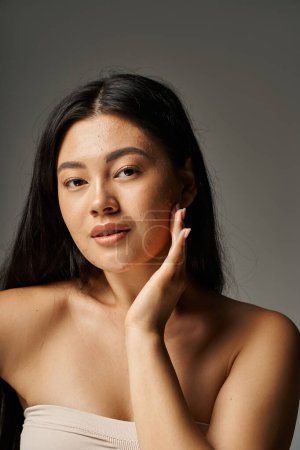vertical view of young asian woman with skin issues and bare shoulders looking at camera on grey